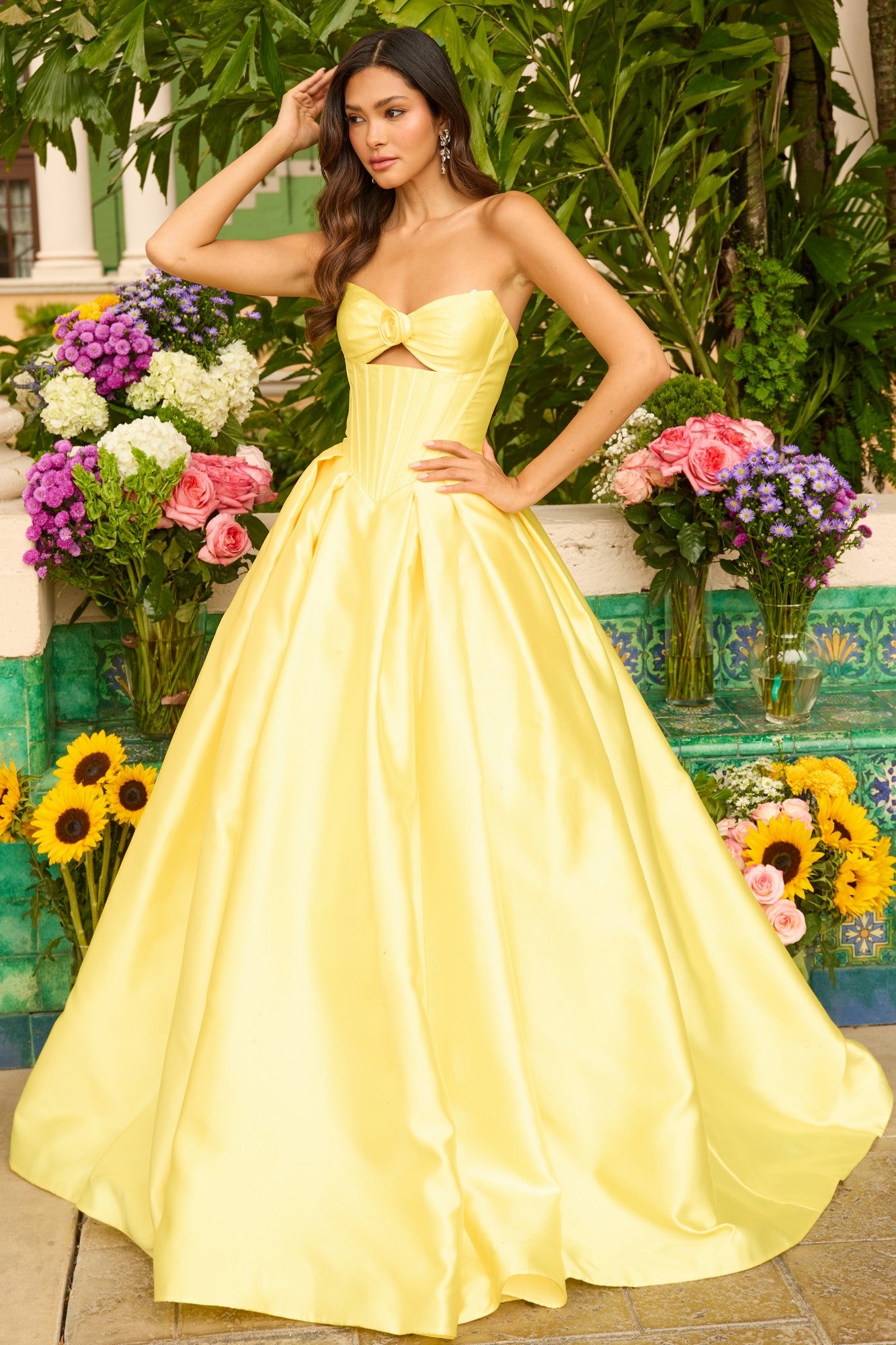 A woman wearing the Ava Presley 47223 Strapless Satin Prom Ball Gown stands outdoors, surrounded by vibrant flower arrangements and lush greenery.